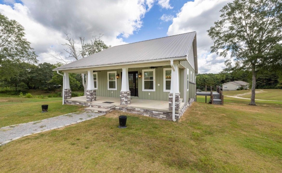 White house with a dark porch sits beside a road lined with trees in autumn colors.
