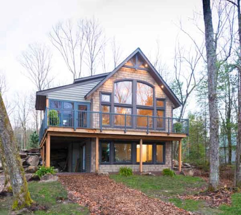 Two-story cabin with wood siding, large arched windows, and a balcony in a wooded setting.