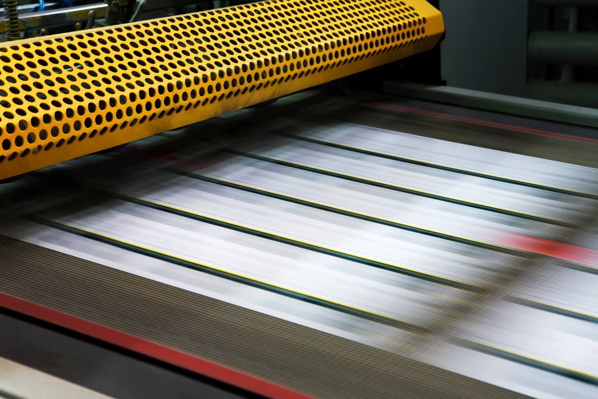Yellow safety guard over a conveyor belt carrying metal strips, industrial setting.