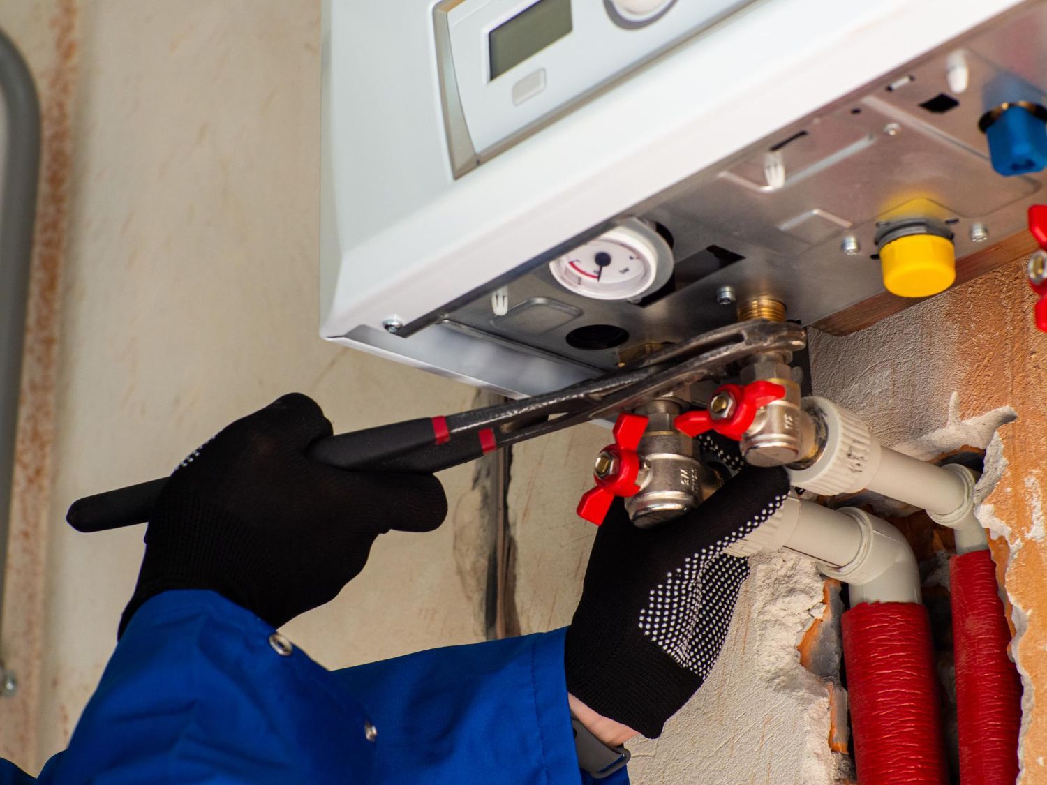 Technician adjusting pipes under a wall-mounted boiler with a wrench, surrounded by red insulated plumbing lines.