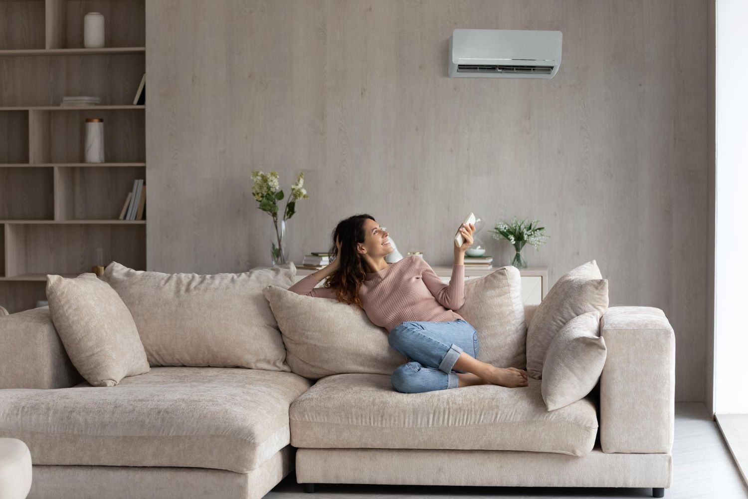 Woman relaxing on a beige sofa in a minimalist living room, holding a drink and looking at her phone