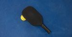 Black pickleball paddle with a yellow perforated ball on a blue surface