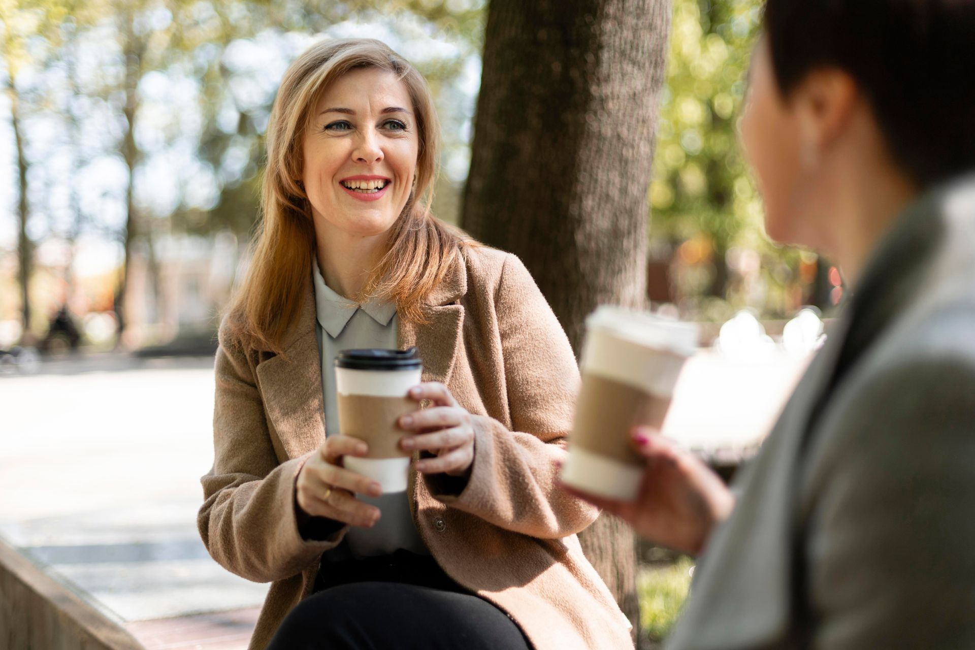 Two women are sitting under a tree holding cups of coffee.