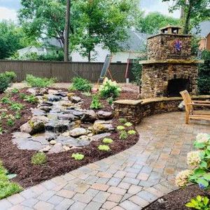 A stone fireplace and waterfall feature stand in a backyard with a brick patio, mulch beds, and surrounding trees.