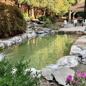 A stone-lined garden pond with a waterfall, surrounded by trees and a patio with chairs in a backyard.