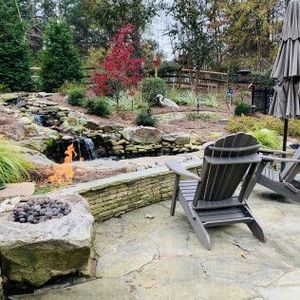 A stone patio with a fire pit and Adirondack chairs overlooking a landscaped backyard featuring a small waterfall.