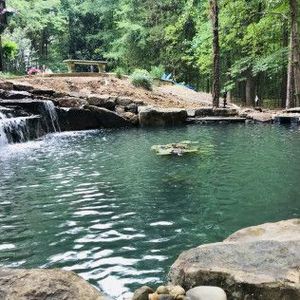 A small waterfall flows into a calm, green swimming pond surrounded by rocky edges, lush trees, and a wooden bench.