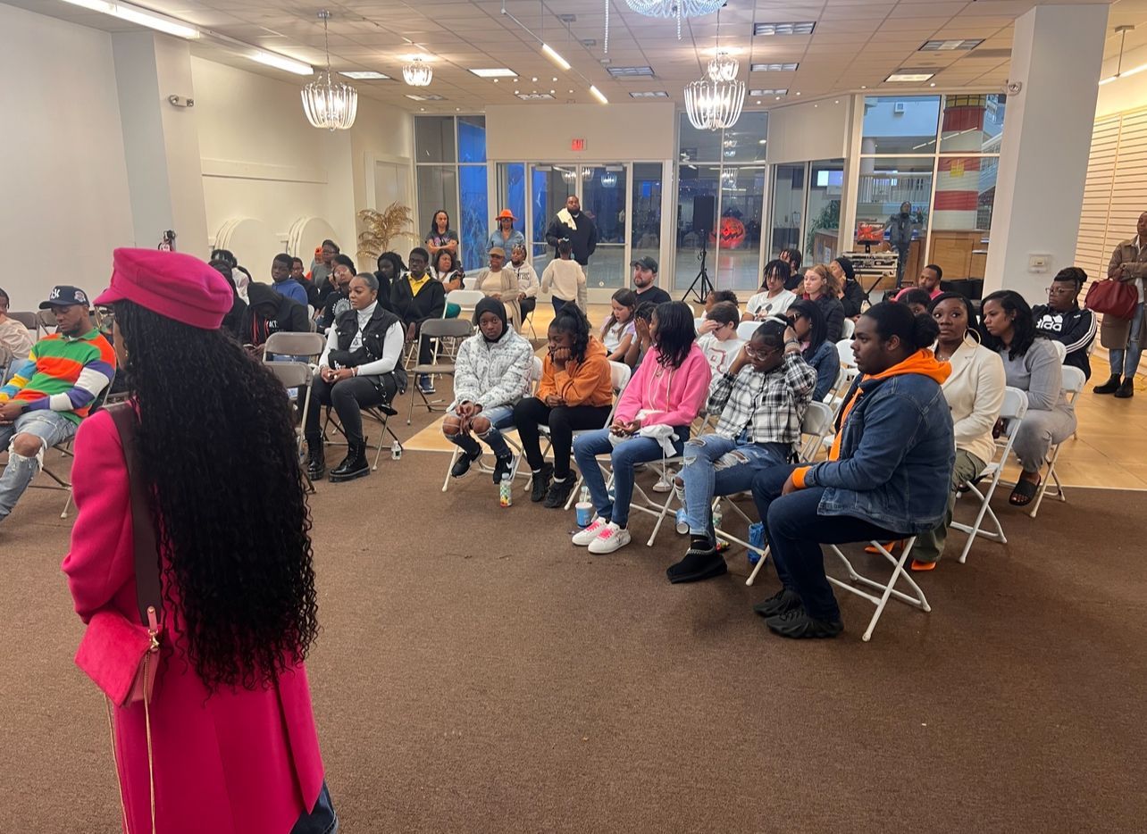 A woman in a pink coat is standing in front of a group of people sitting in chairs.