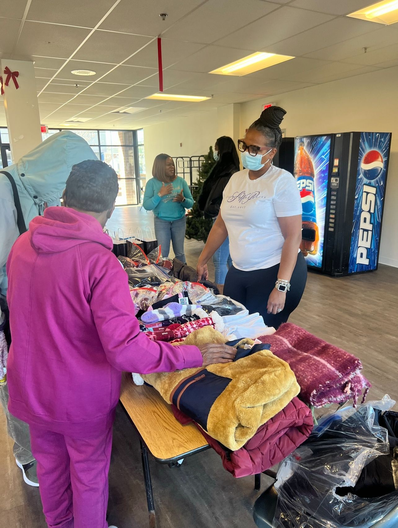 A group of people are standing around a table with blankets and a pepsi machine in the background.