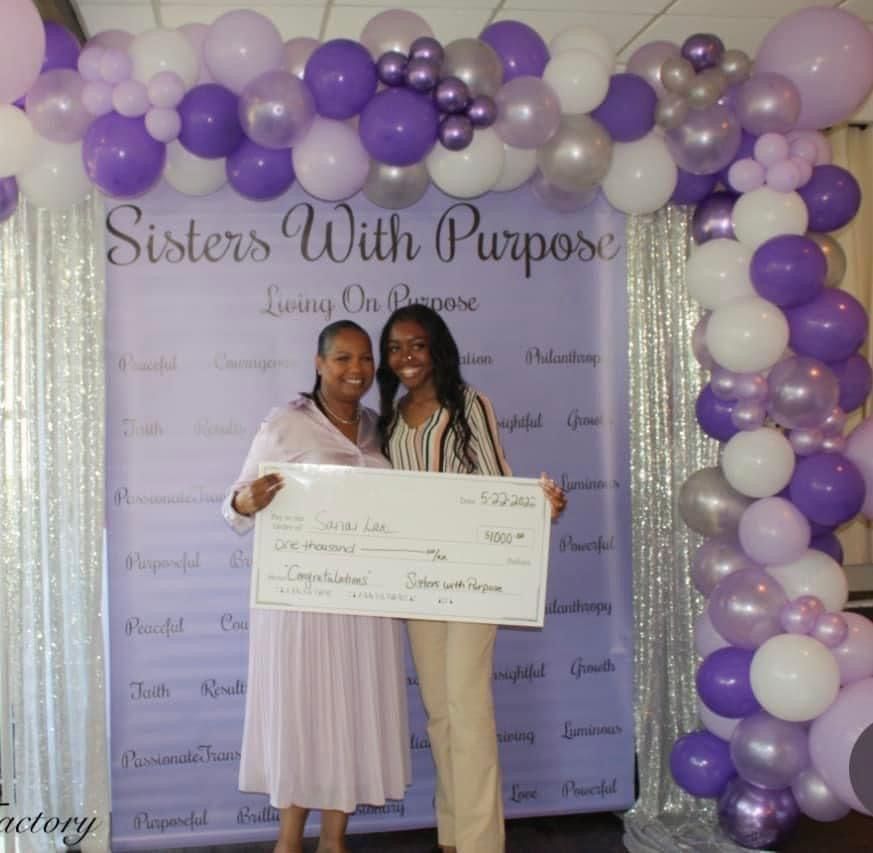 Two women holding a check in front of a sign that says sisters with purpose