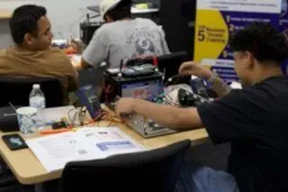 People working at a cluttered table with electronics and papers in an indoor workshop.