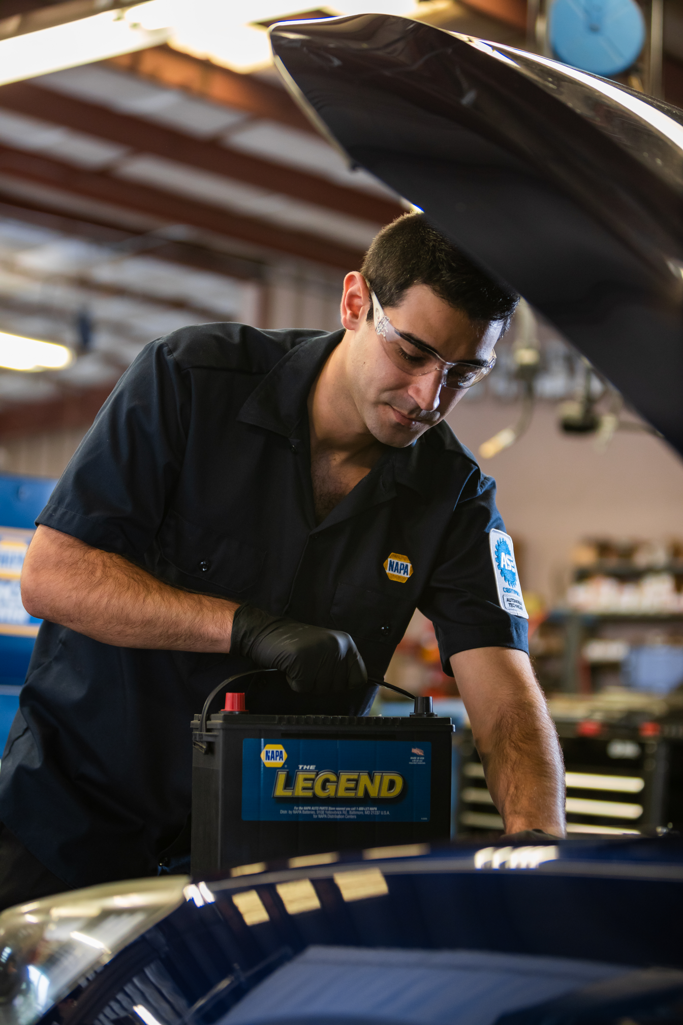 Mechanic working under an open car hood with a battery charger in an auto repair shop