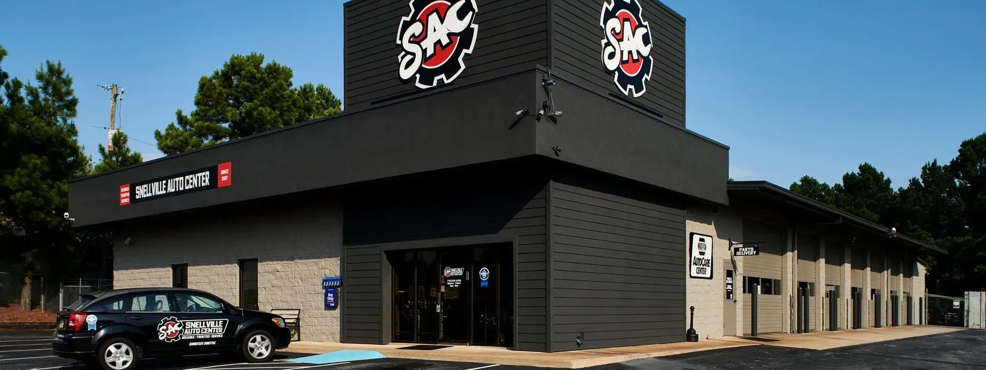 Exterior of a black storefront with red logo, large windows, and parked cars on a sunny day