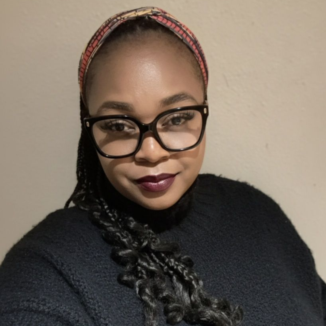 Woman with glasses, wearing a dark sweater and patterned headband. She is smiling slightly, with dark lipstick.
