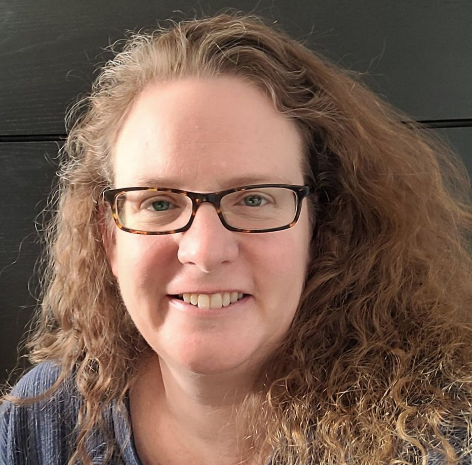 Woman with glasses, wearing a dark sweater and patterned headband. She is smiling slightly, with dark lipstick.