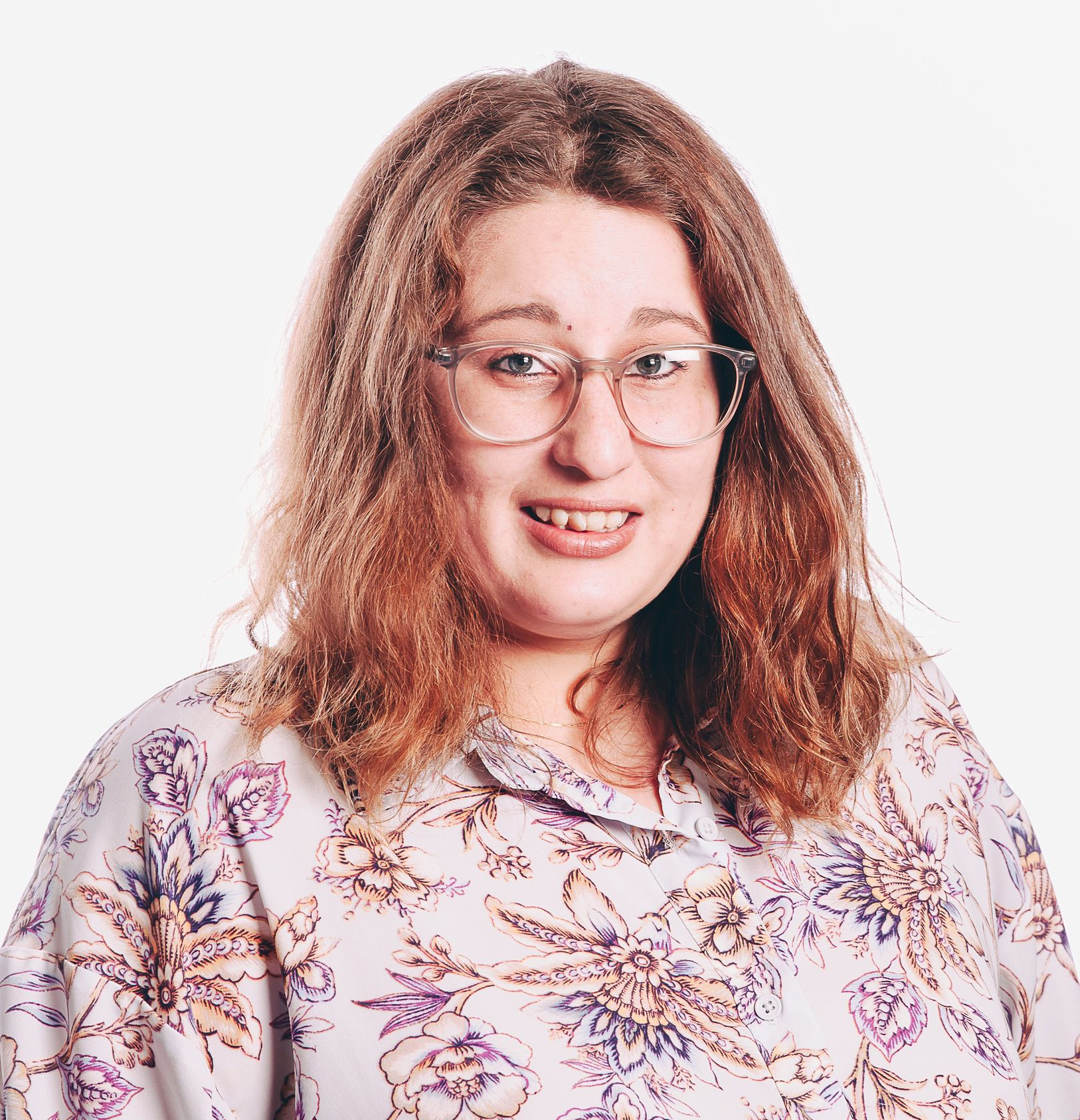 Woman with curly auburn hair, glasses, and floral shirt smiling at the camera. Studio setting.