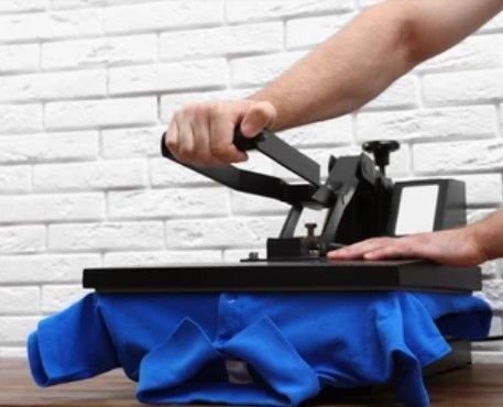 Person using a heat press machine to apply a design to a blue t-shirt on a wooden table against a white brick wall.