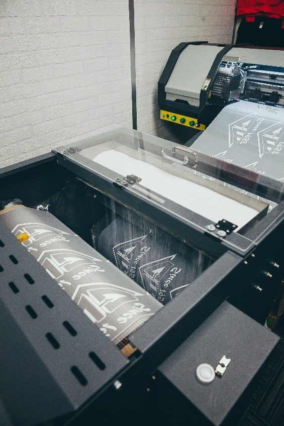 Large format printer with roll of printed material inside. Black machine with clear cover and grey print.