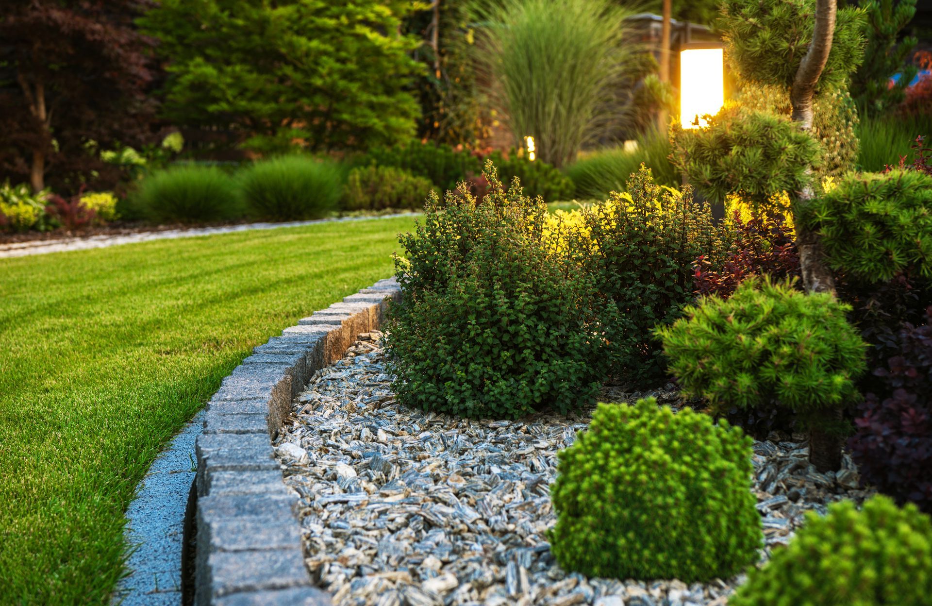 A well-manicured lawn with a stone-bordered gravel garden bed containing small shrubs, illuminated by a garden light.