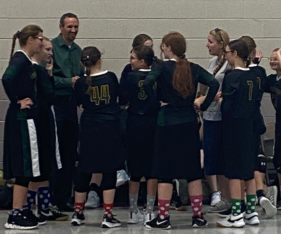 Indoor basketball game with players in green and black jerseys under a red hoop