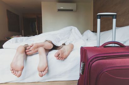 Dos personas durmiendo en una cama, con los pies visibles bajo las sábanas blancas y una maleta roja en primer plano.