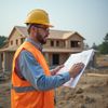 Construction worker in hard hat and vest reviews blueprints at a house under construction.