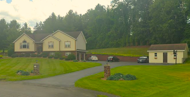 House with cars in the driveway, small outbuilding, and grassy lawn in a wooded area.