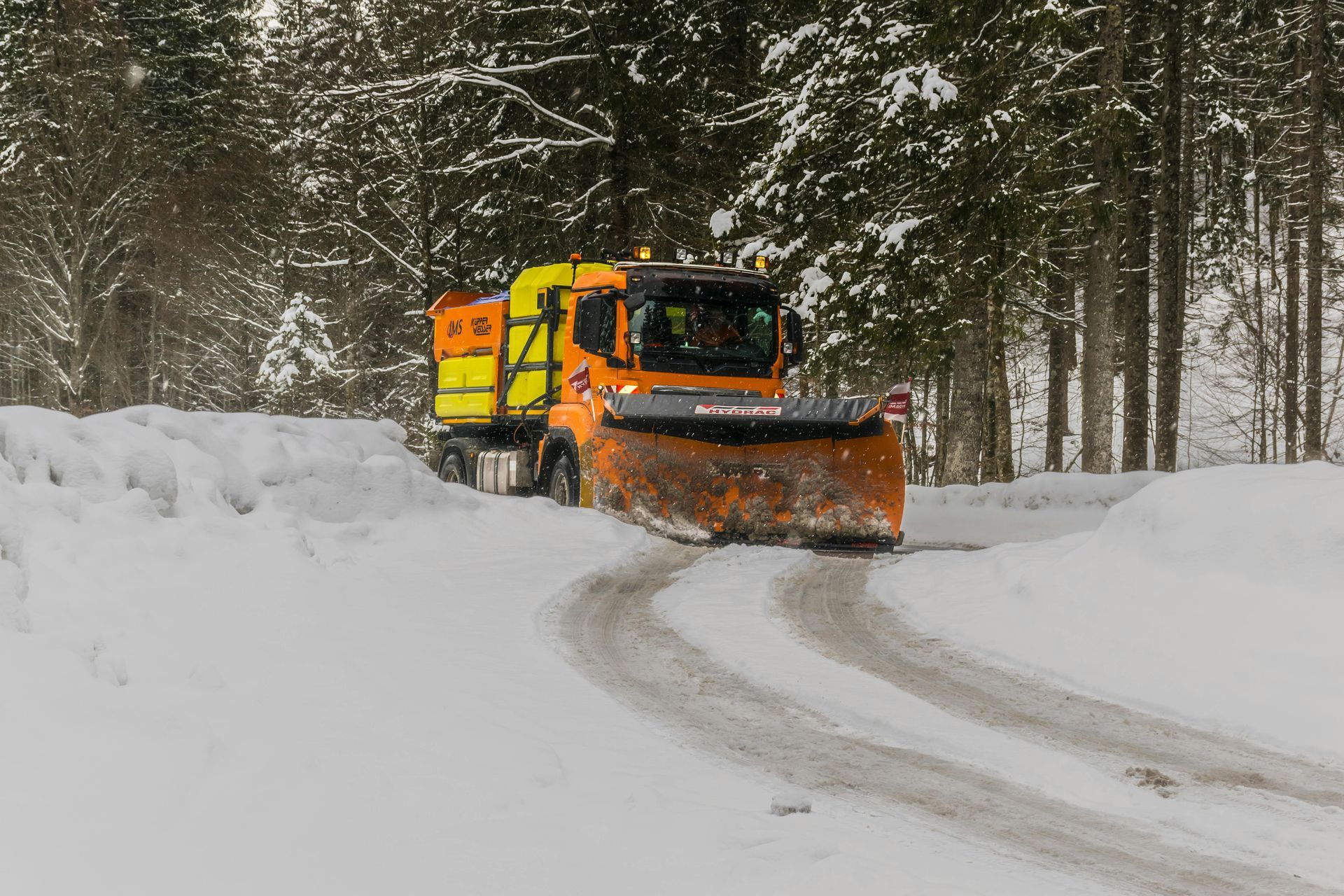 Orange snowplow clearing a snow-covered road in a wooded area.