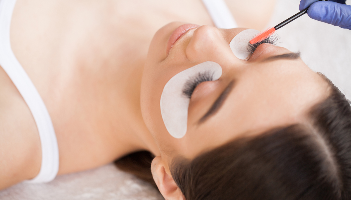Woman with eye patches receiving eyelash extensions; technician using a brush.
