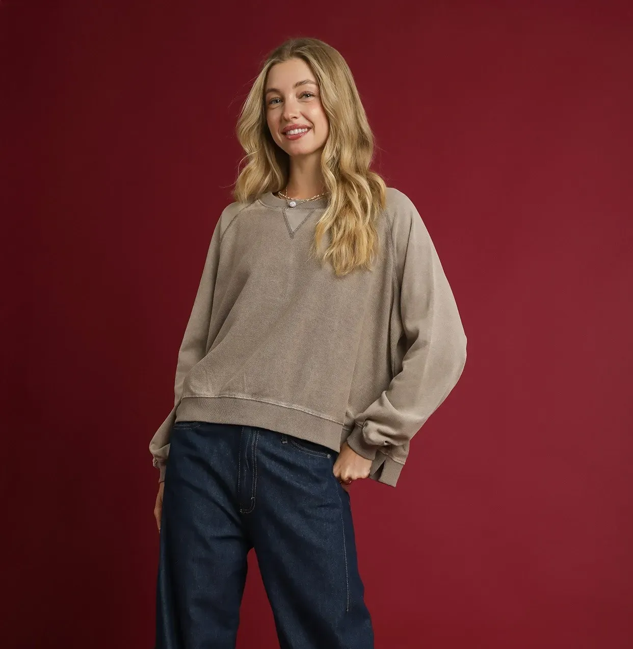 A woman in a brown sweatshirt and blue jeans smiles, poses against a red background.