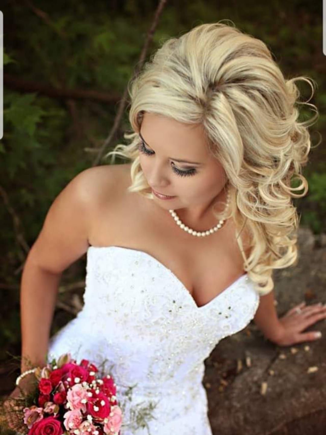 Bride in a white beaded strapless wedding dress, looking down, holding a bouquet of red and pink flowers, with curly blonde hair.