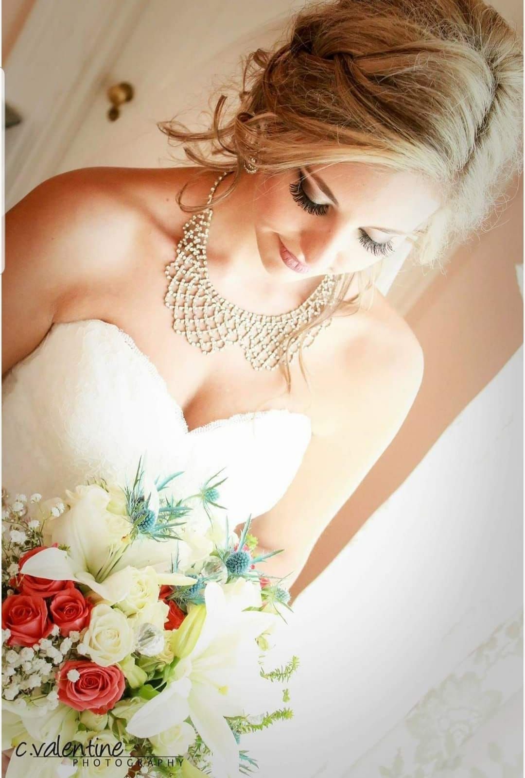 Bride in a white strapless gown holding bouquet, looking down. Updo hairstyle, ornate necklace.