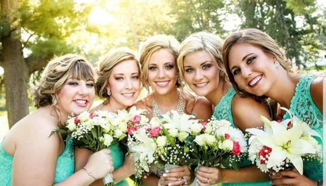Five women smiling, holding bouquets, wearing teal dresses, outdoors in sunlight.