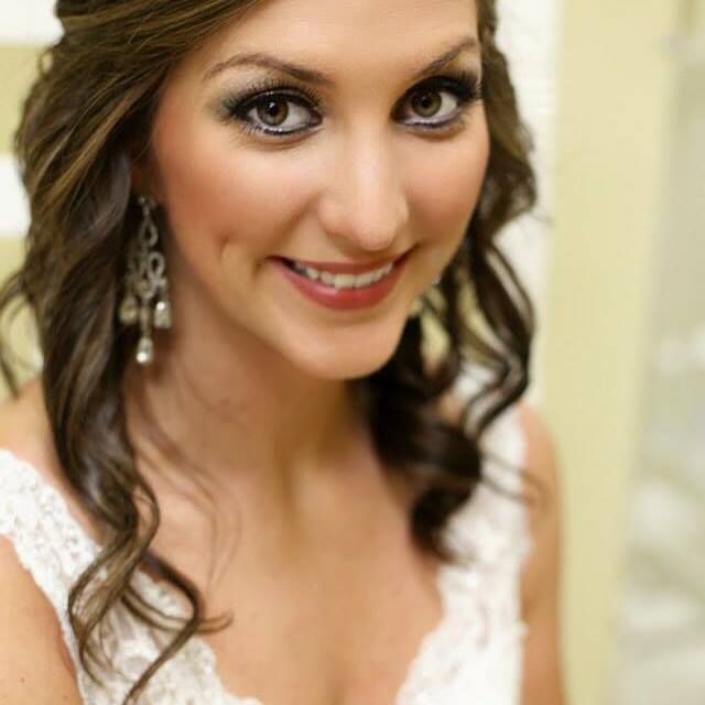 Woman with brown wavy hair, wearing drop earrings, and a white lace dress smiles.