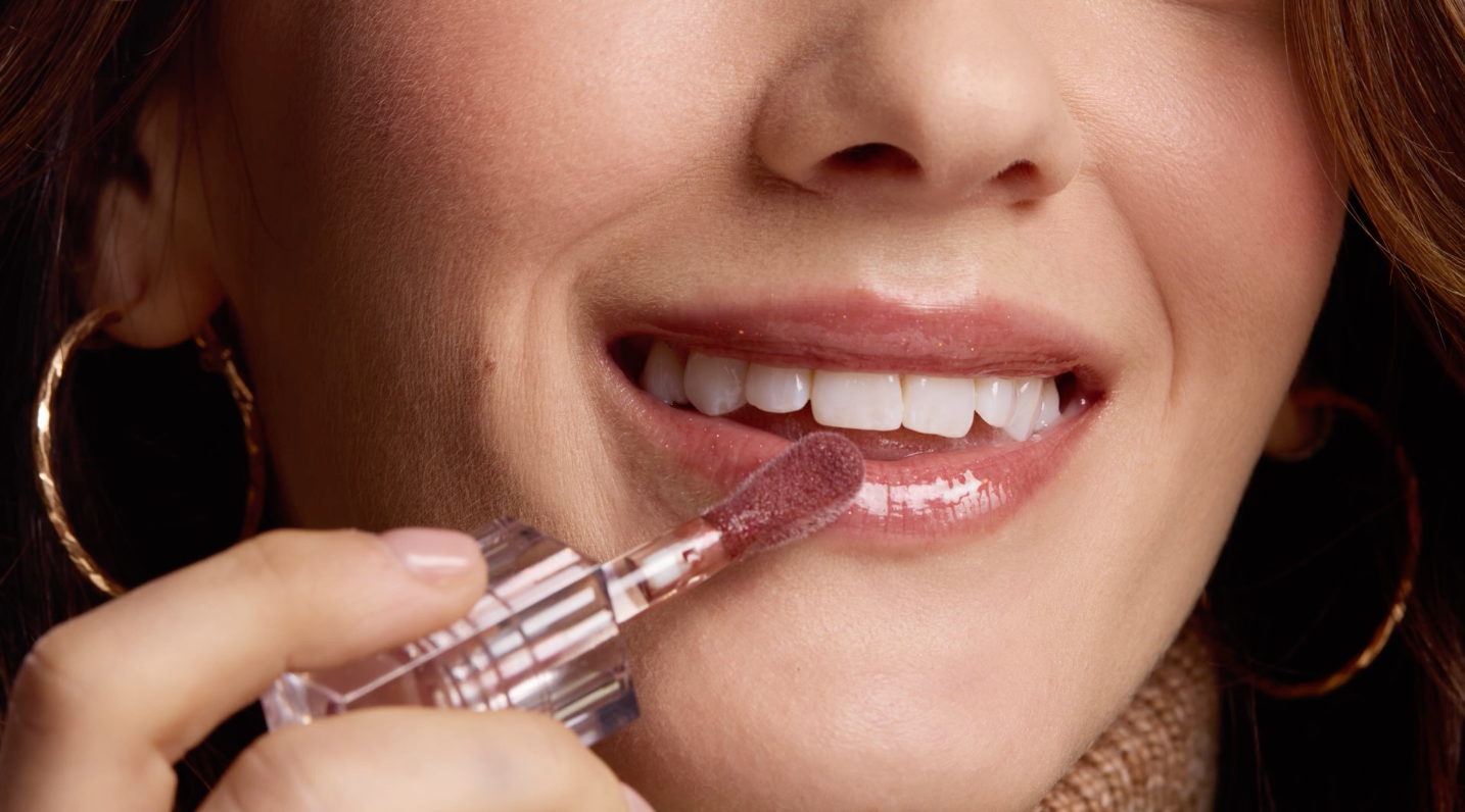 Woman applying lip gloss; smiling, indoors.