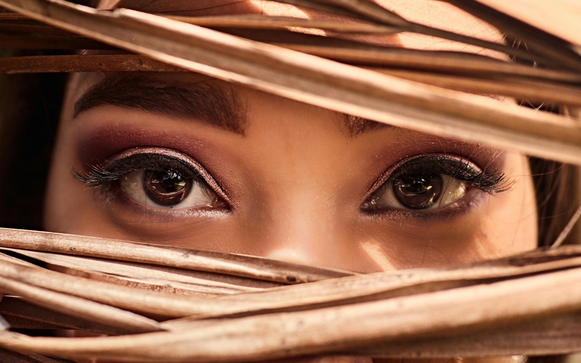 Eyes peeking through brown foliage. Dark eyeshadow, long lashes, focused gaze.