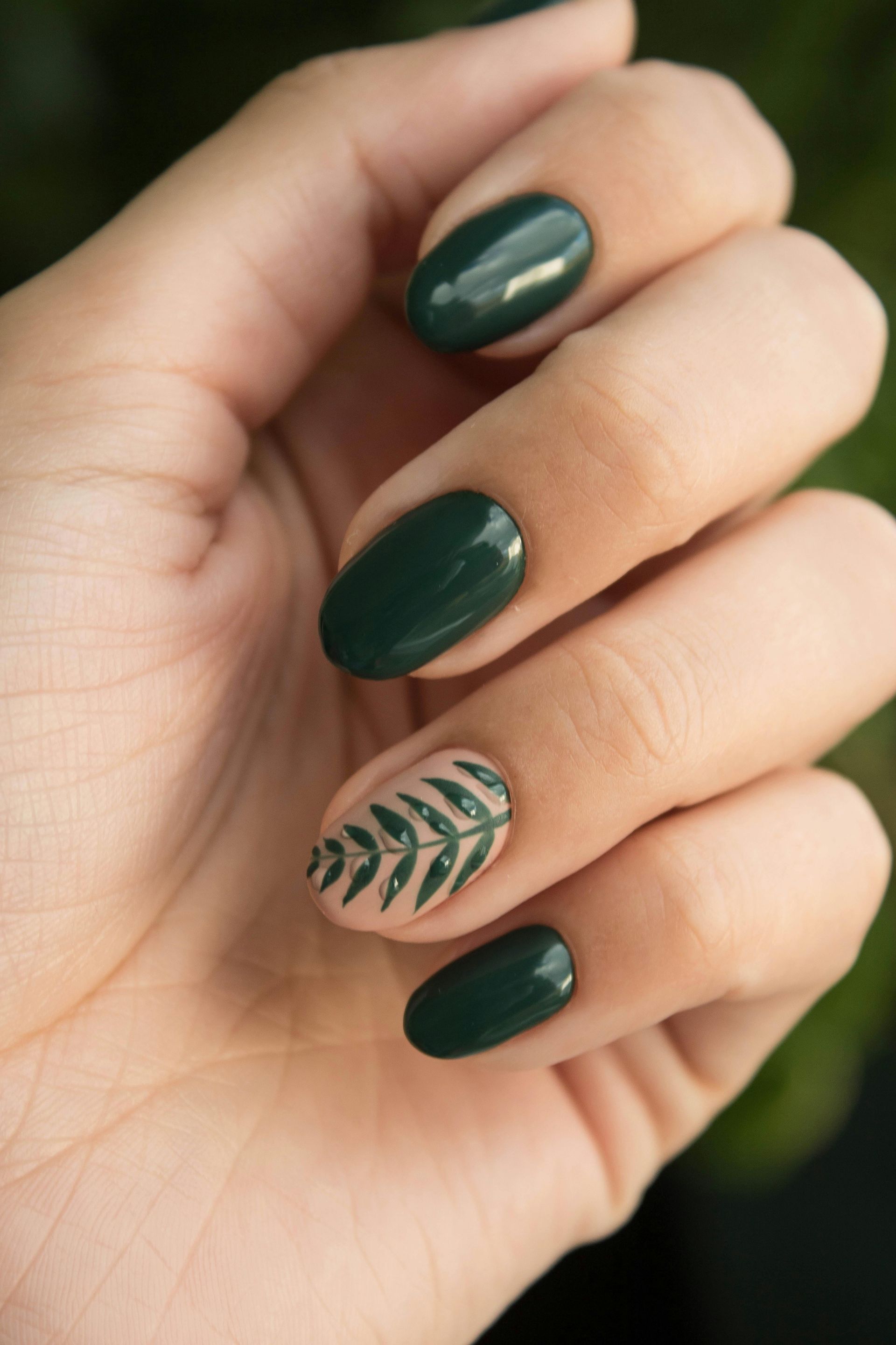 Hand with dark green polished nails; one nail features a beige leaf design.