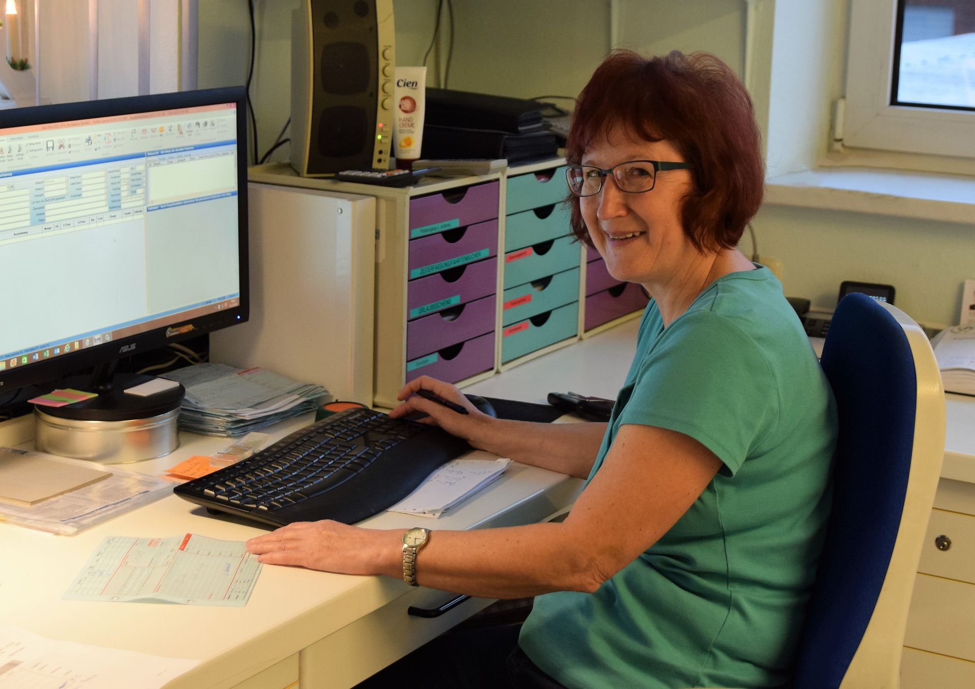 Frau mit Brille lächelt, während sie in einem Büro an einem Computer arbeitet und eine Tastatur benutzt.