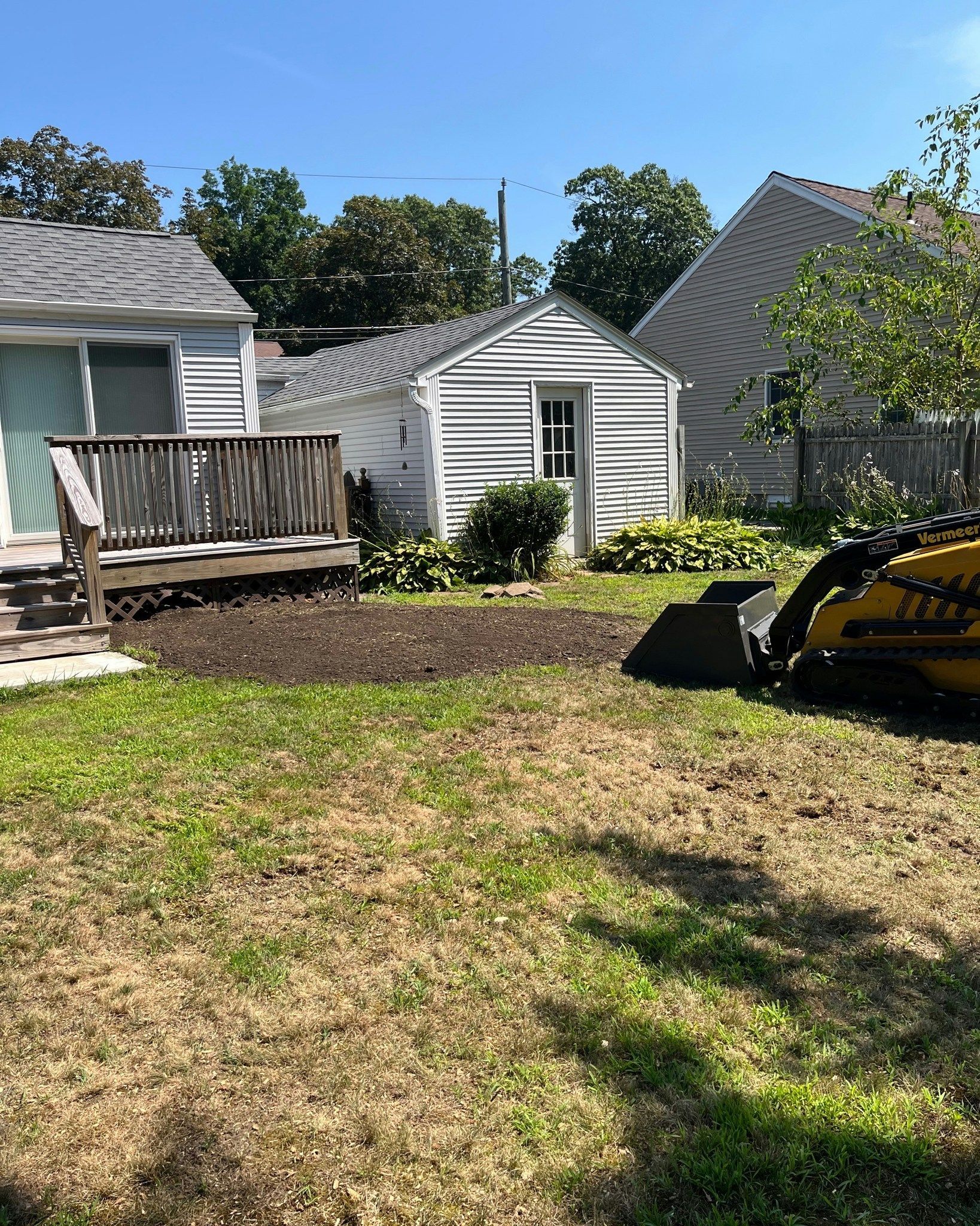 Backyard with a small shed and deck; recently tilled dark soil in the center of the frame. Yellow excavator on the right.