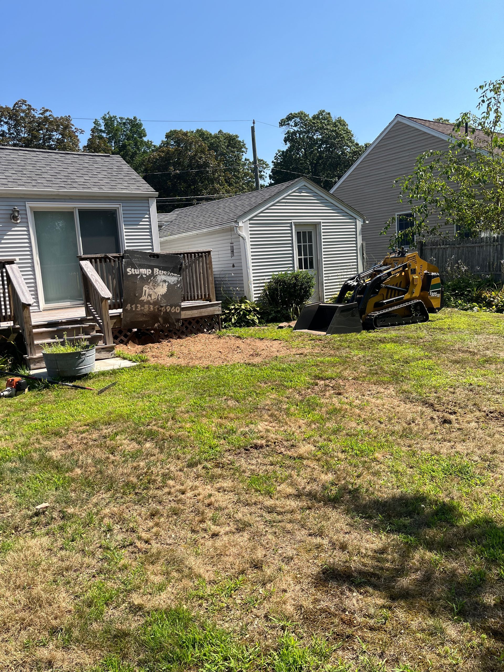 A small backyard with a stump grinder next to a small shed. Brown woodchips scattered on the ground.