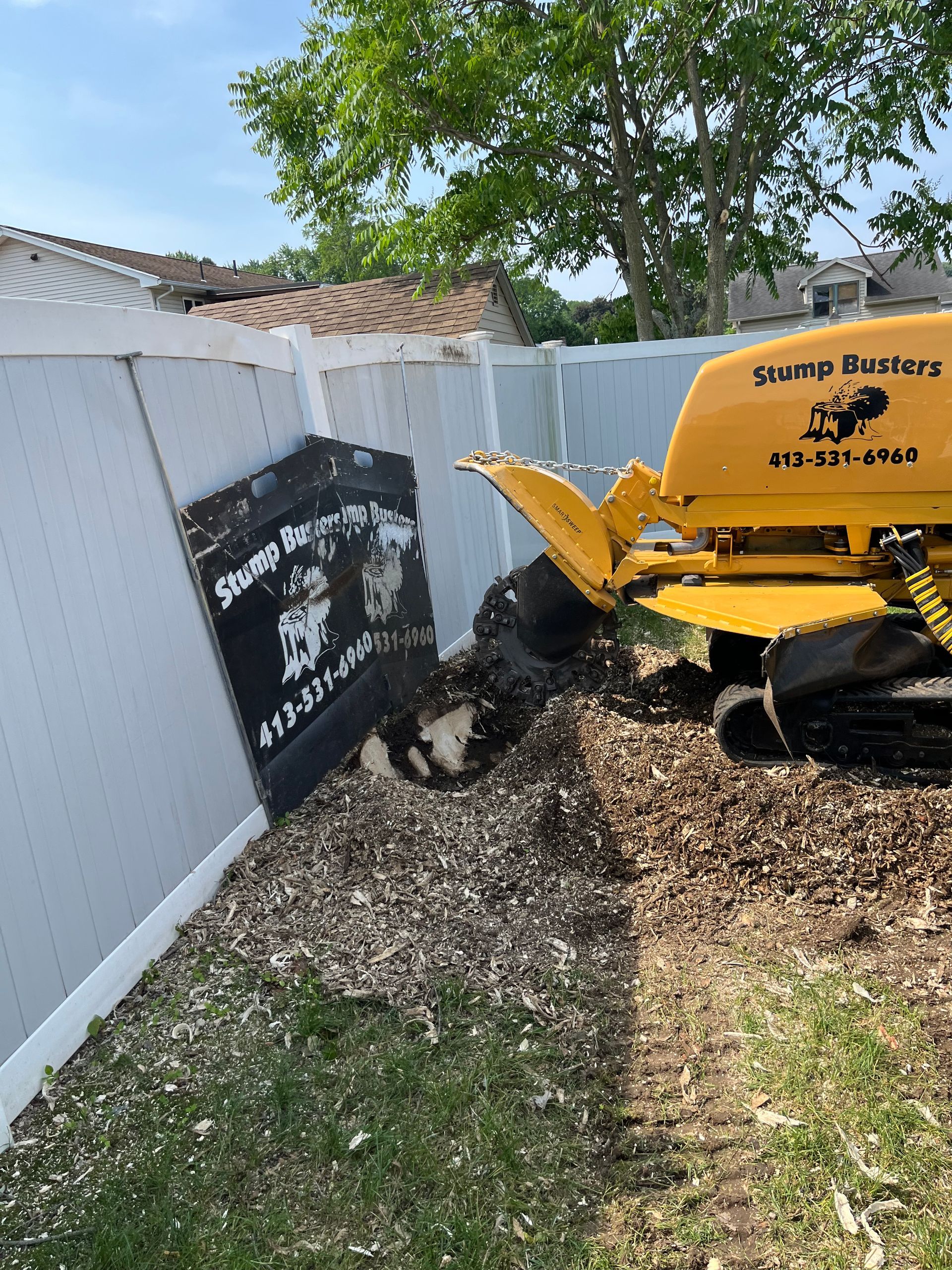 Yellow stump grinder grinding a tree stump near a white fence, creating wood chips on the ground.