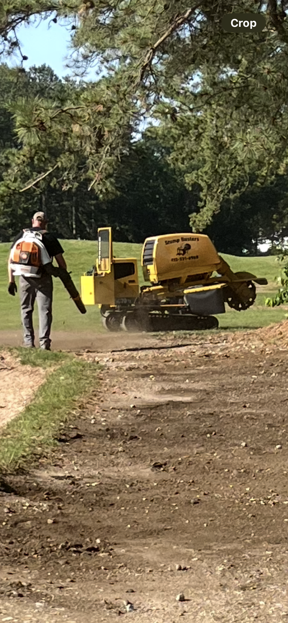 A worker using a leaf blower near a yellow stump grinder on a dirt path, trees in the background.