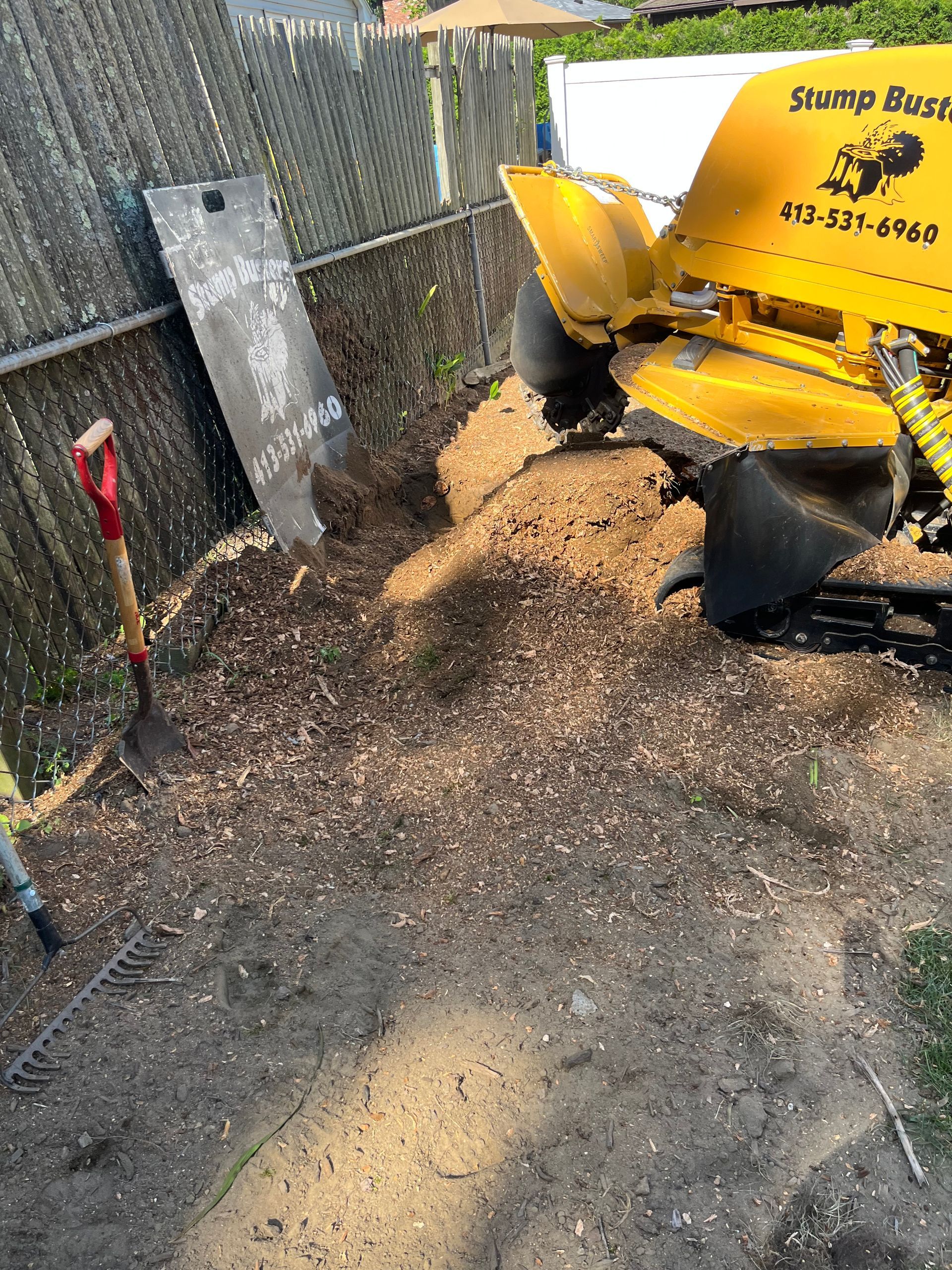 A yellow stump grinder working next to a wooden fence, grinding wood chips. A shovel rests nearby.