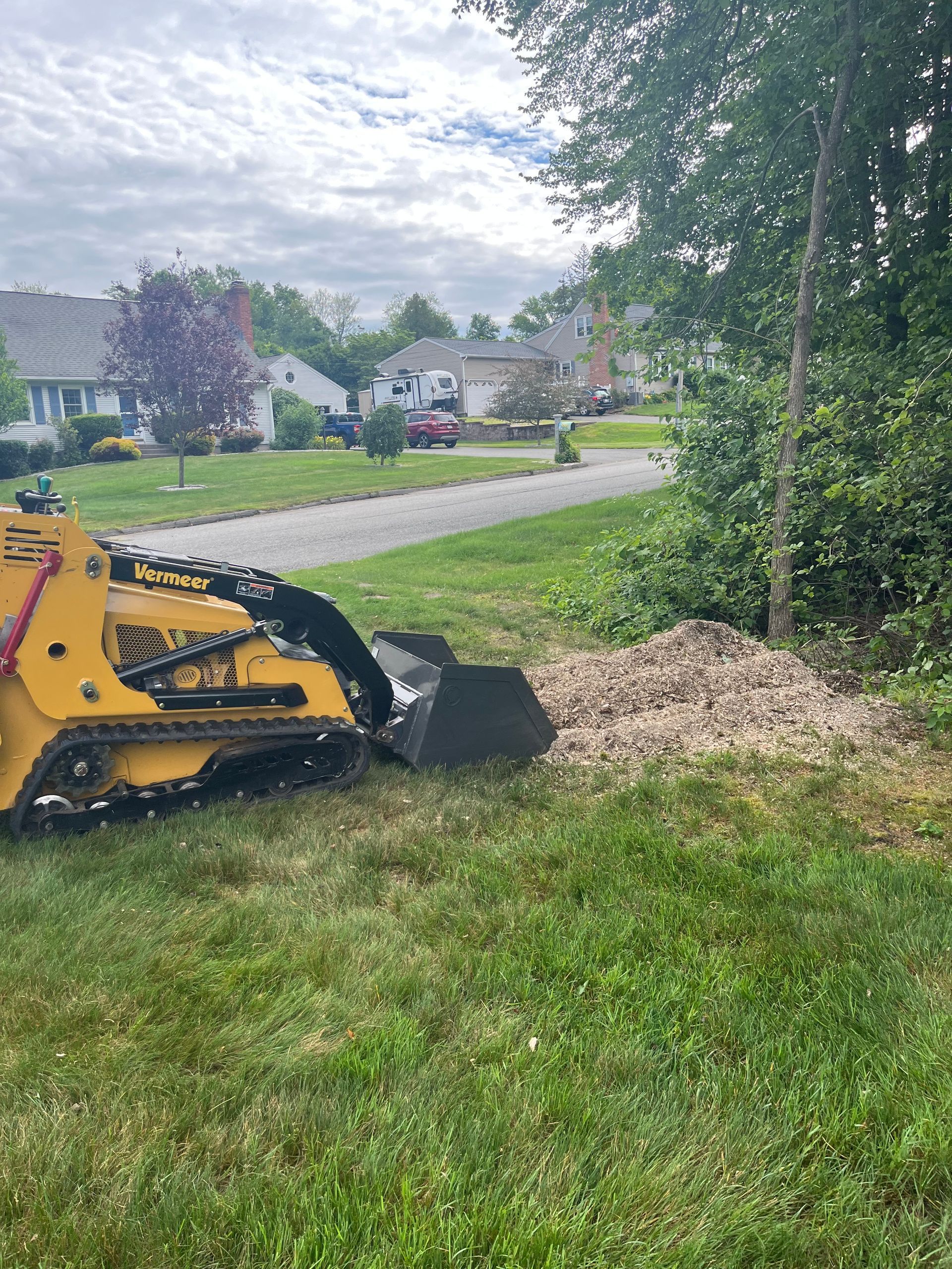 Yellow skid steer with bucket moving gravel on a grassy area next to a road, with houses in the background.