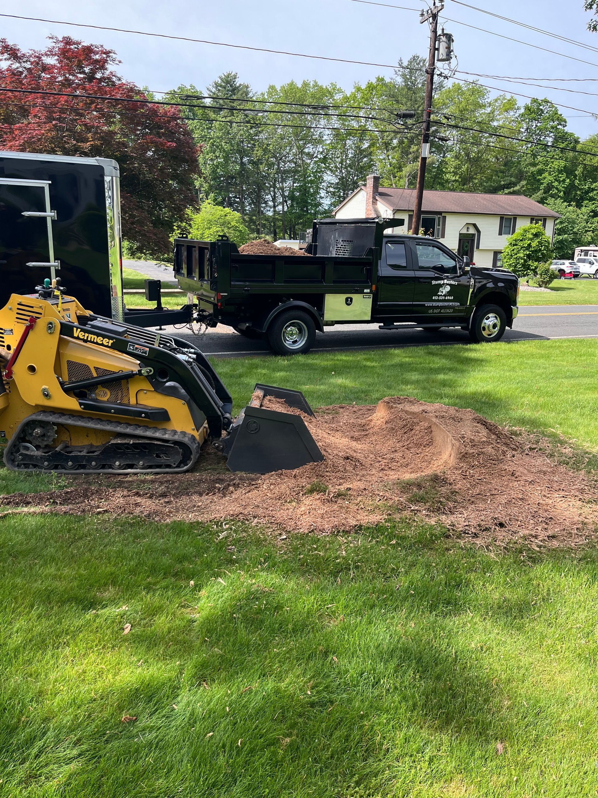Yellow skid steer loading dirt into a black dump truck and trailer on a lawn.