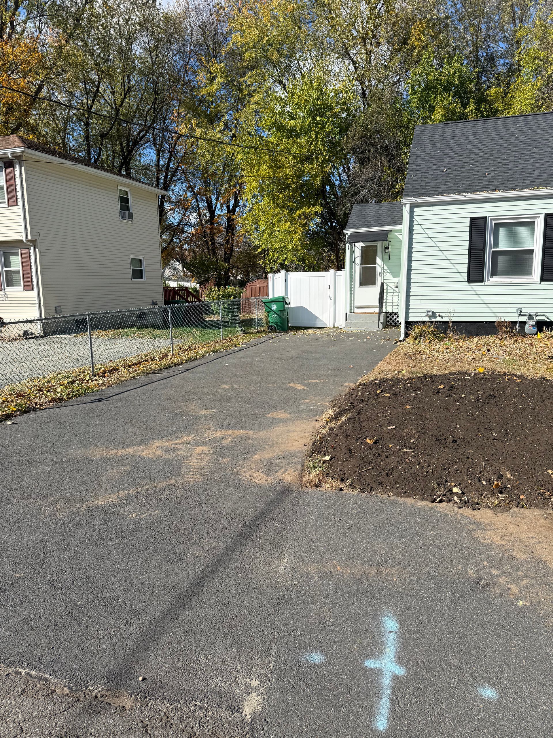 Driveway leading to a small house with black asphalt. Dirt pile and chain-link fence on the sides; fall foliage visible.