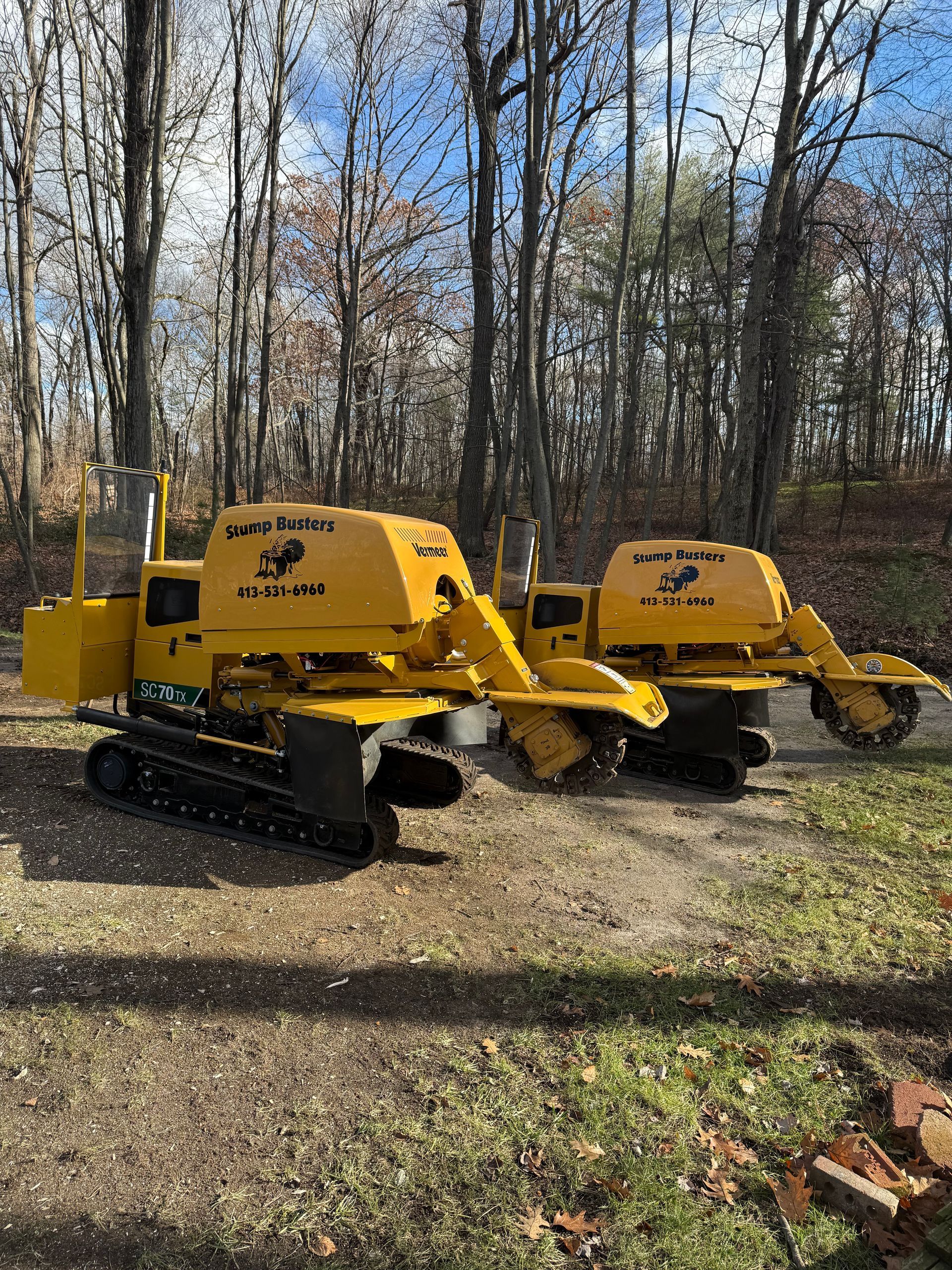 Two yellow stump grinders on tracks parked on a gravel path near trees.