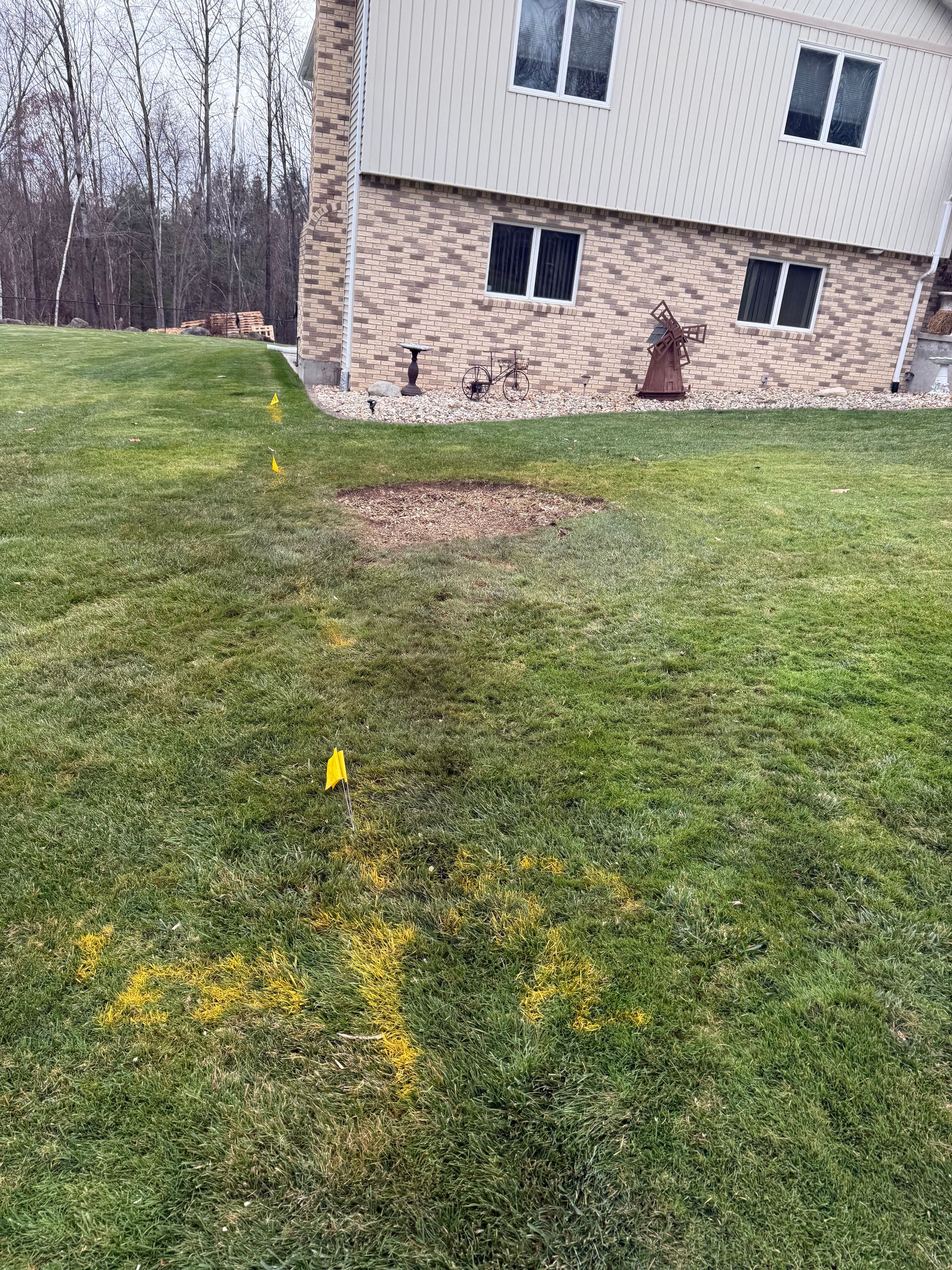 Yellow spray paint and flags mark buried utilities on a lawn near a two-story brick house.