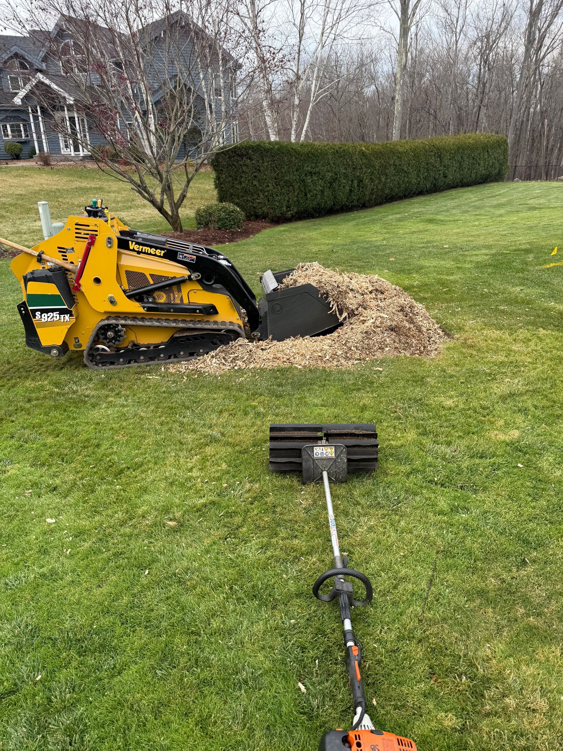 Yellow skid steer digging hole in a green lawn next to a string trimmer.