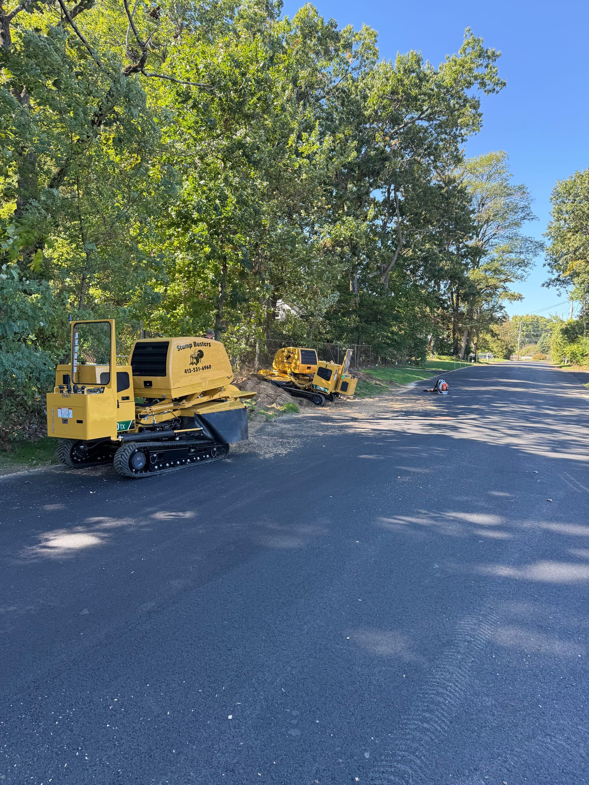 Yellow stump grinder on a paved road, grinding wood near trees. Sunny day.